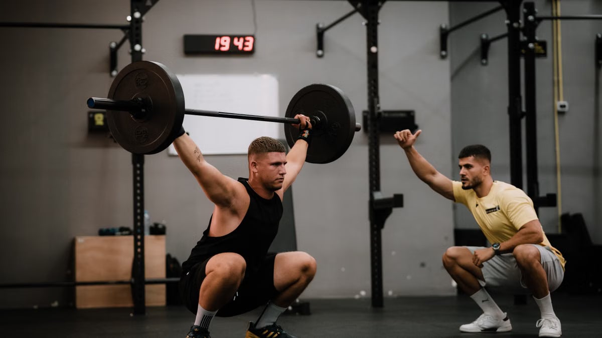 Muscular man lifting a barbell in a gym under his trainer's guidance, showcasing strength and focus.